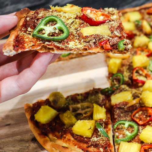 A hand holding a Pineapple Pepper Plant-based Pizza slice