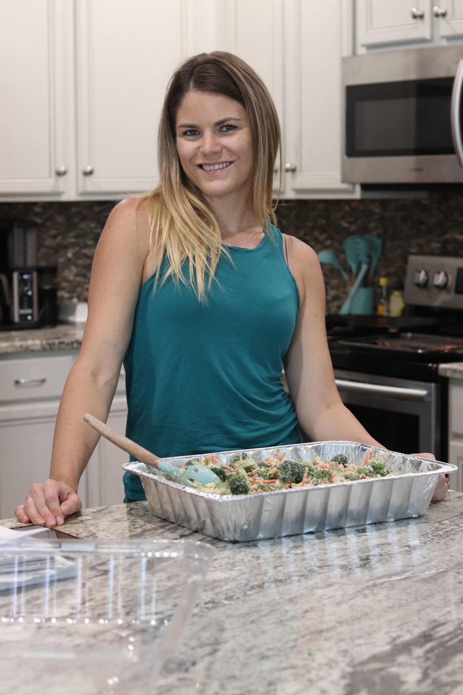Caitlin Havener Rogers smiling in her kitchen behind a large foil pan of broccoli salad